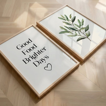Two framed art prints displayed on a light wood herringbone floor. One print features the text 'Good Food Brighter Days' in black serif font with a small heart, and the other features a green leafy branch illustration.