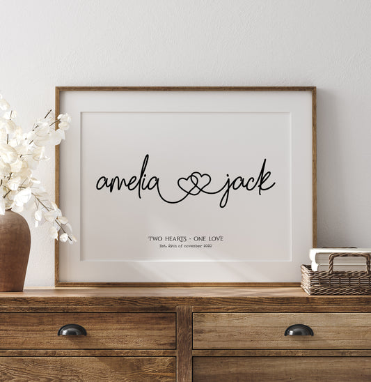 A personalised names print illustrating two names written in a fancy font joined by two hearts in a continuous line. Underneath are the words 'Two hearts, one love' and the couples marriage date in a black font.