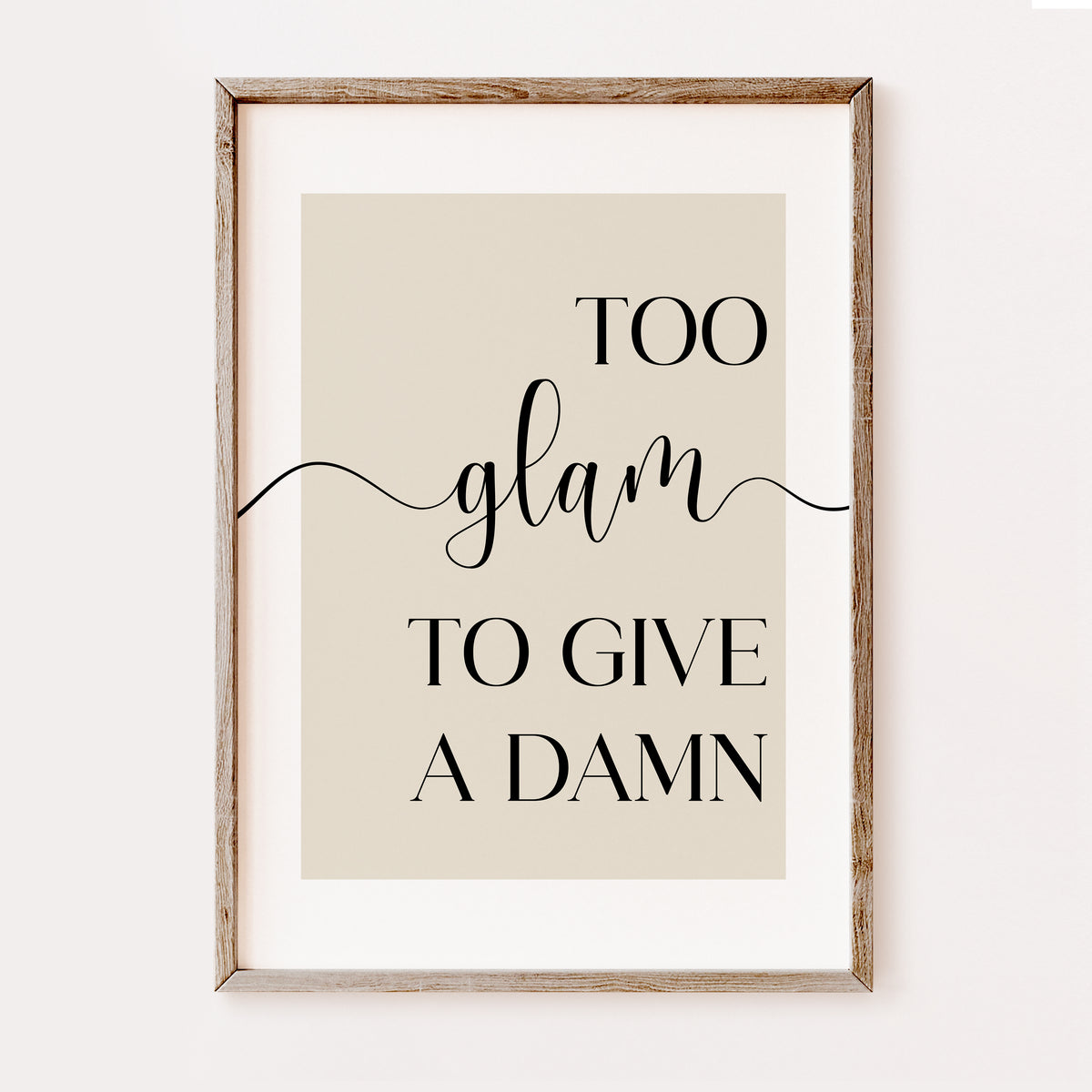 Framed typographic wall art with the phrase “TOO glam TO GIVE A DAMN” in black lettering on a warm beige background, shown in a light natural wood frame against a white wall.
