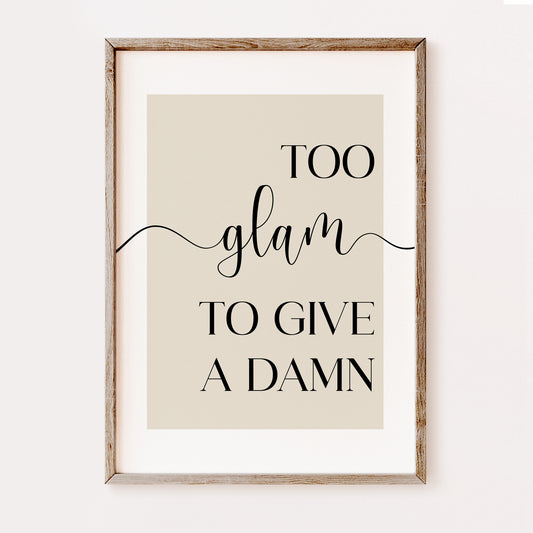 Framed typographic wall art with the phrase “TOO glam TO GIVE A DAMN” in black lettering on a warm beige background, shown in a light natural wood frame against a white wall.